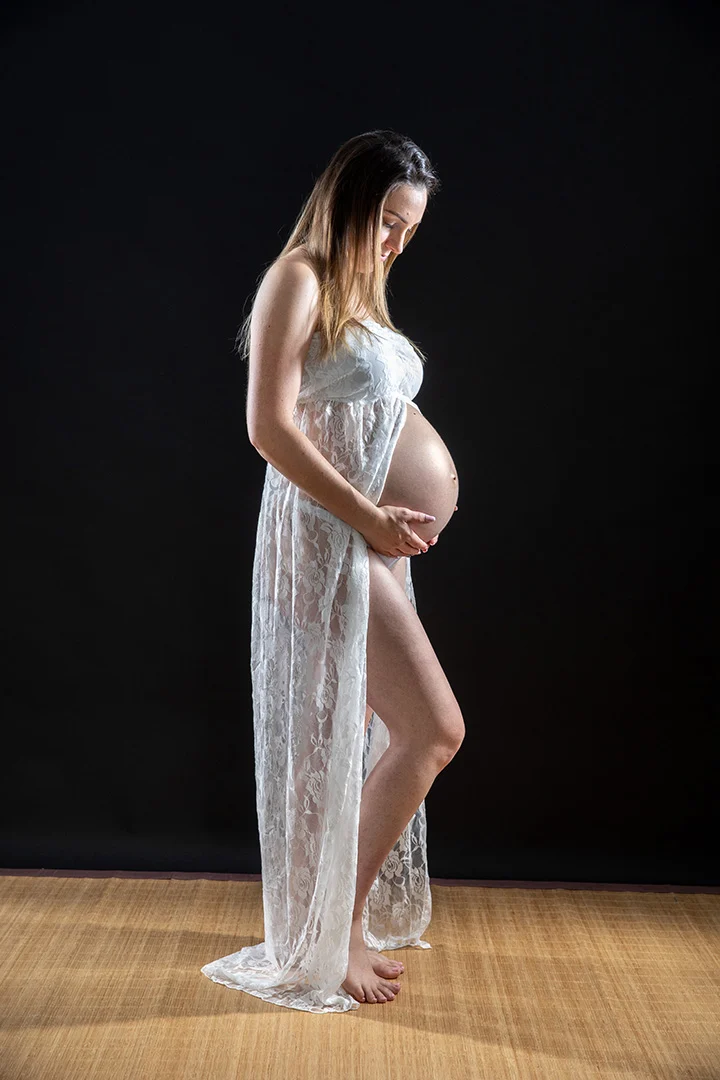 Future maman en robe de dentelle blanche tenant son ventre — séance grossesse en studio
